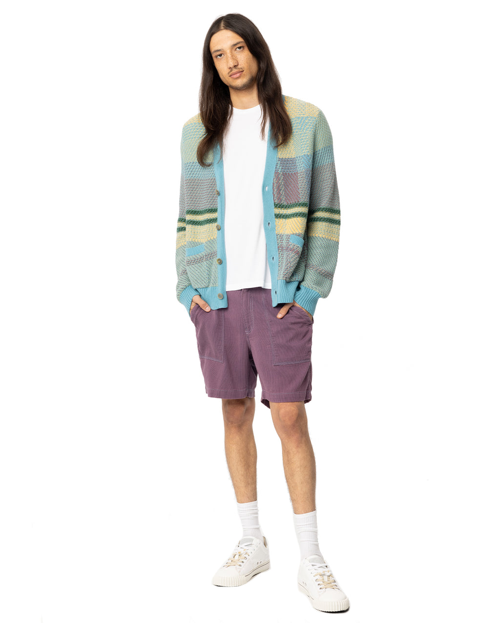 Man standing in purple shorts with a plain white tee under a teal, green and yellow cardigan. 